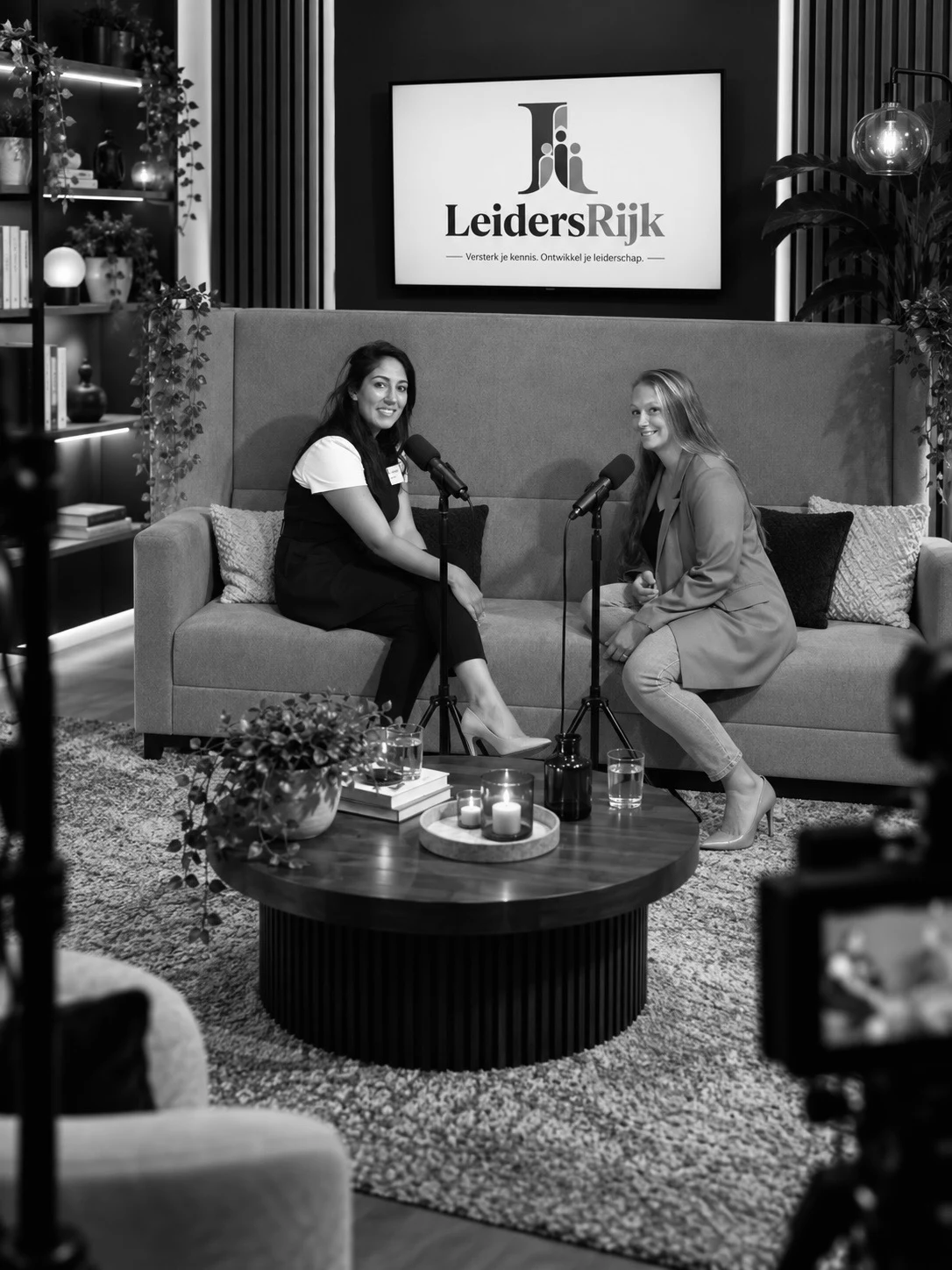 Anisha Biharie en Alice van Leeuwen samen op de bank in de podcaststudio, met het originele LeidersRijk-logo achter hen